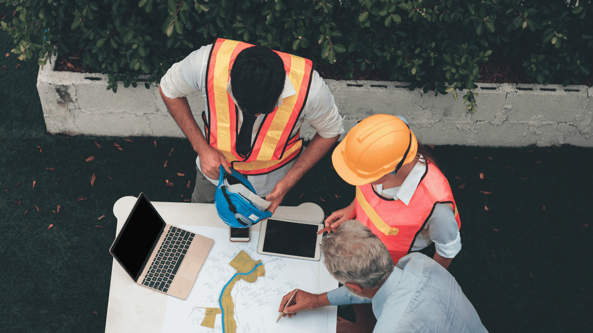 A top down image of three men looking at documents and a laptop on the table. Two men wear hi-visibility vests and one wears a hardhat.